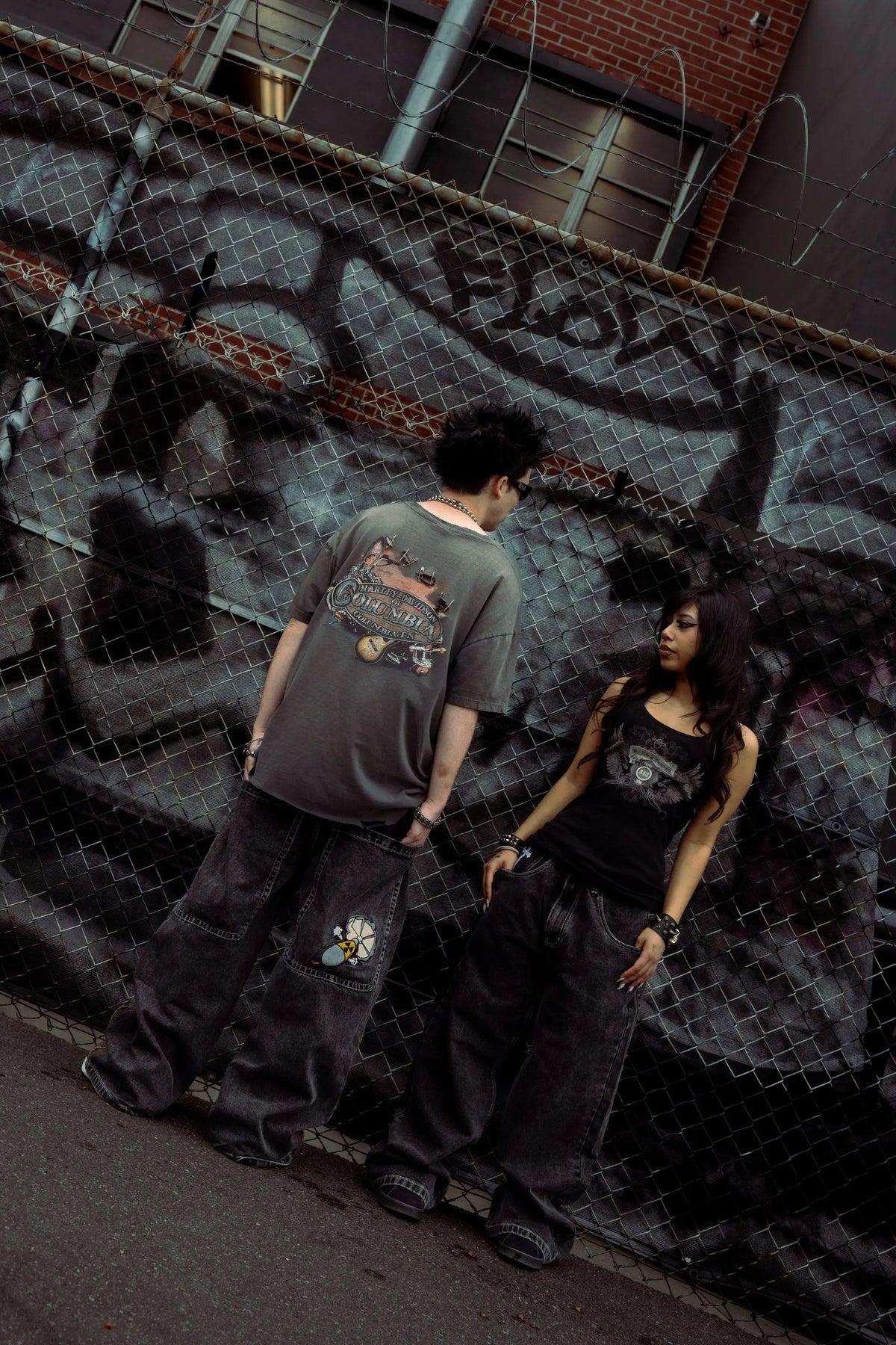 Two models wearing GLOE baggy jeans against a graffiti chain-link fence